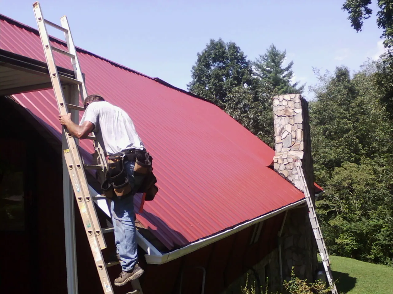 Roofer on a ladder beside a red metal roof and stone chimney in Anna